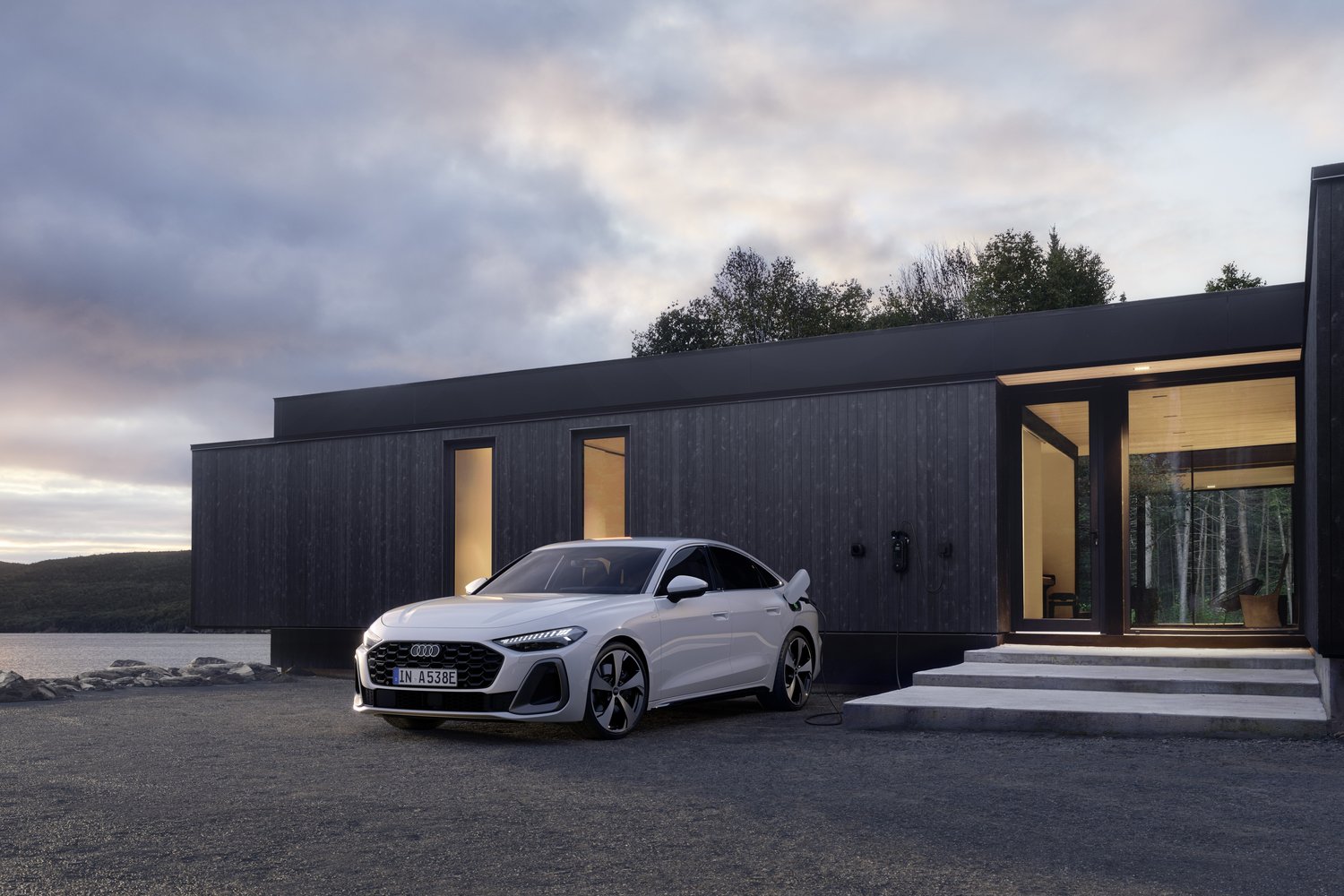 White 2026 Audi A5 e-hybrid quattro plugged in to charge beside a dark timber lakeside cabin