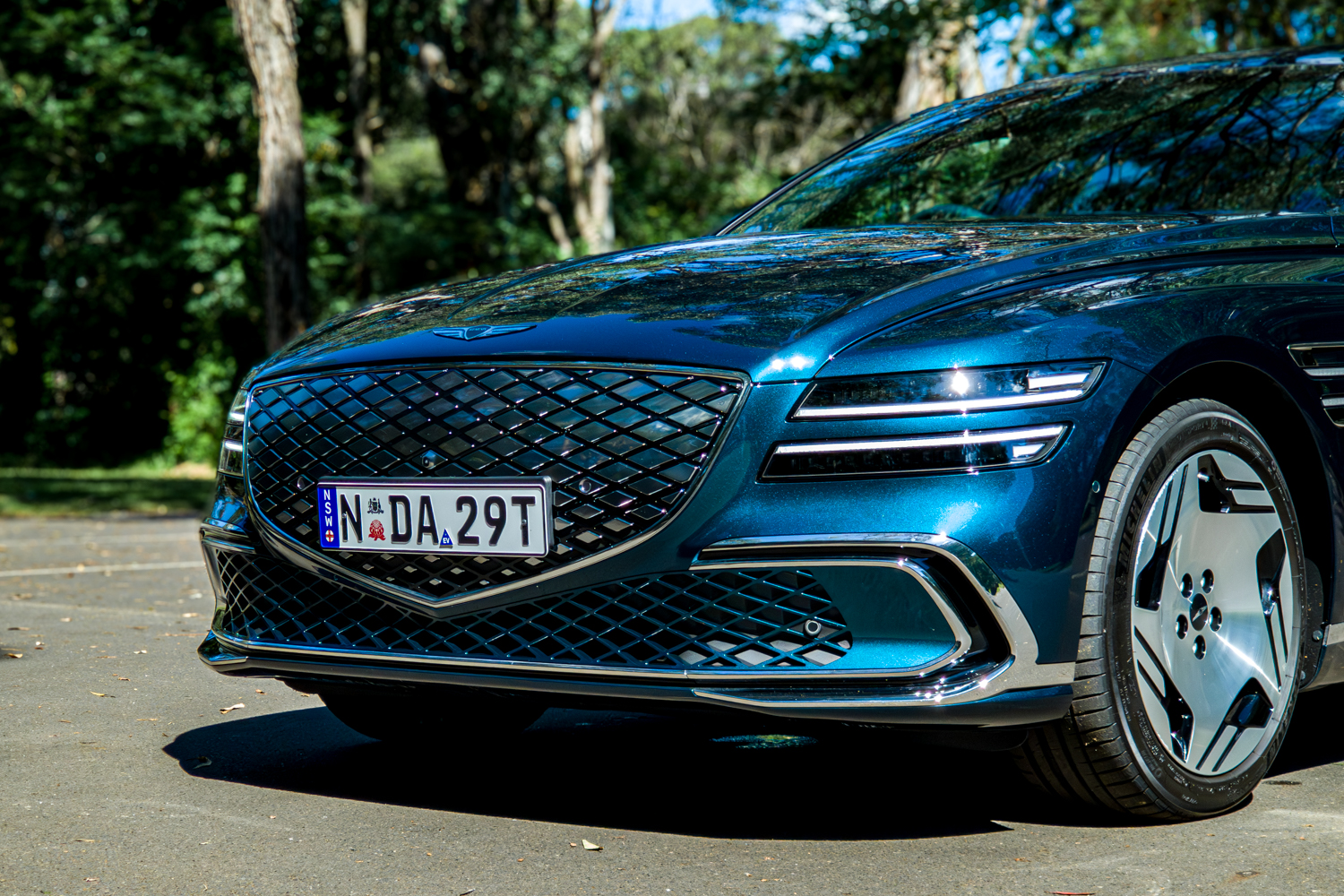 2026 Genesis G80 Electrified Matira Blue front passenger side low angle showing grille headlight and alloy wheel
