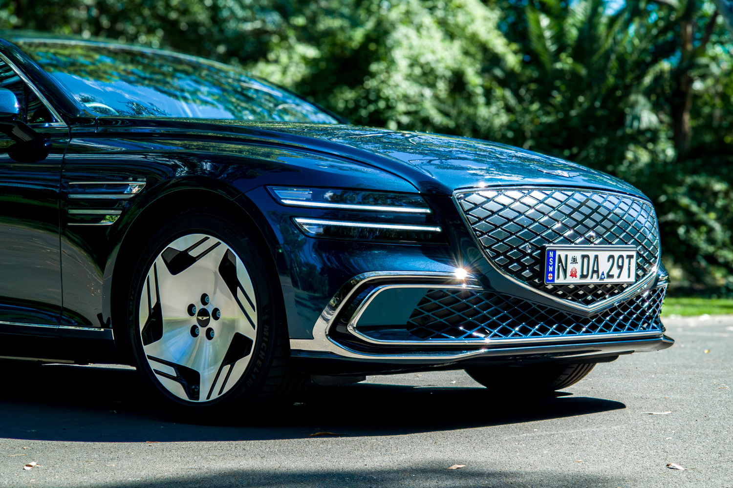 2026 Genesis G80 Electrified Matira Blue front passenger side low angle showing headlight grille and alloy wheel