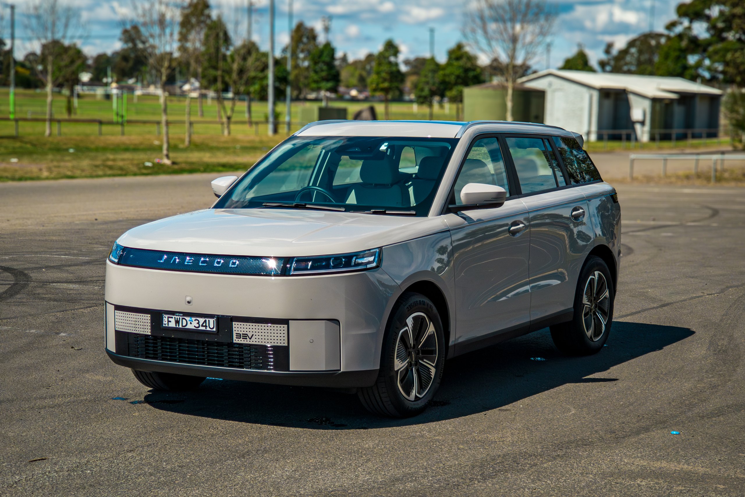 Elevated passenger-side front three-quarter of the Stone Grey 2026 Jaecoo J5 EV Summit parked beside sports fields