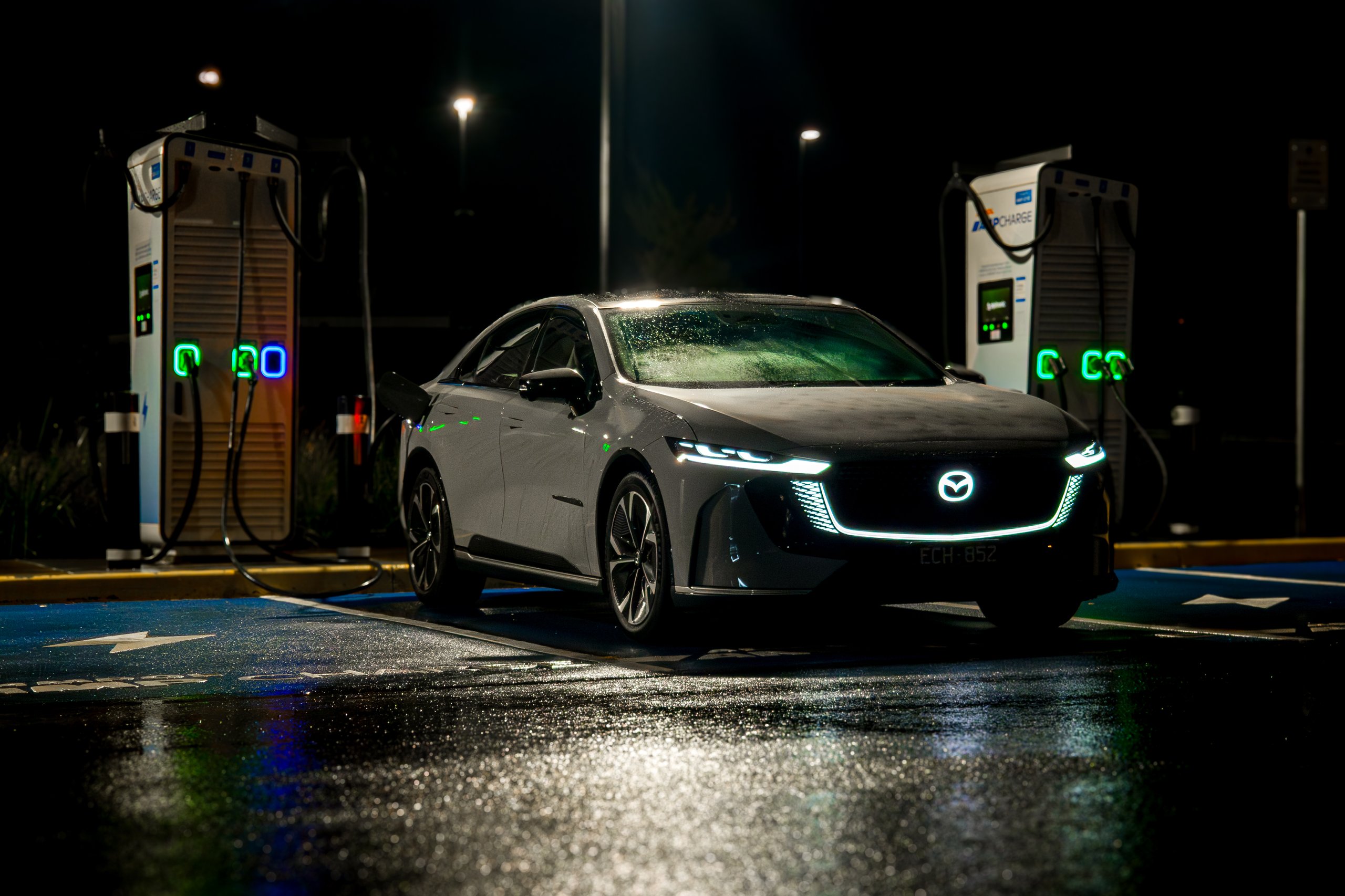 Light grey 2026 Mazda 6e GT plugged into a DC fast charger at night on wet asphalt