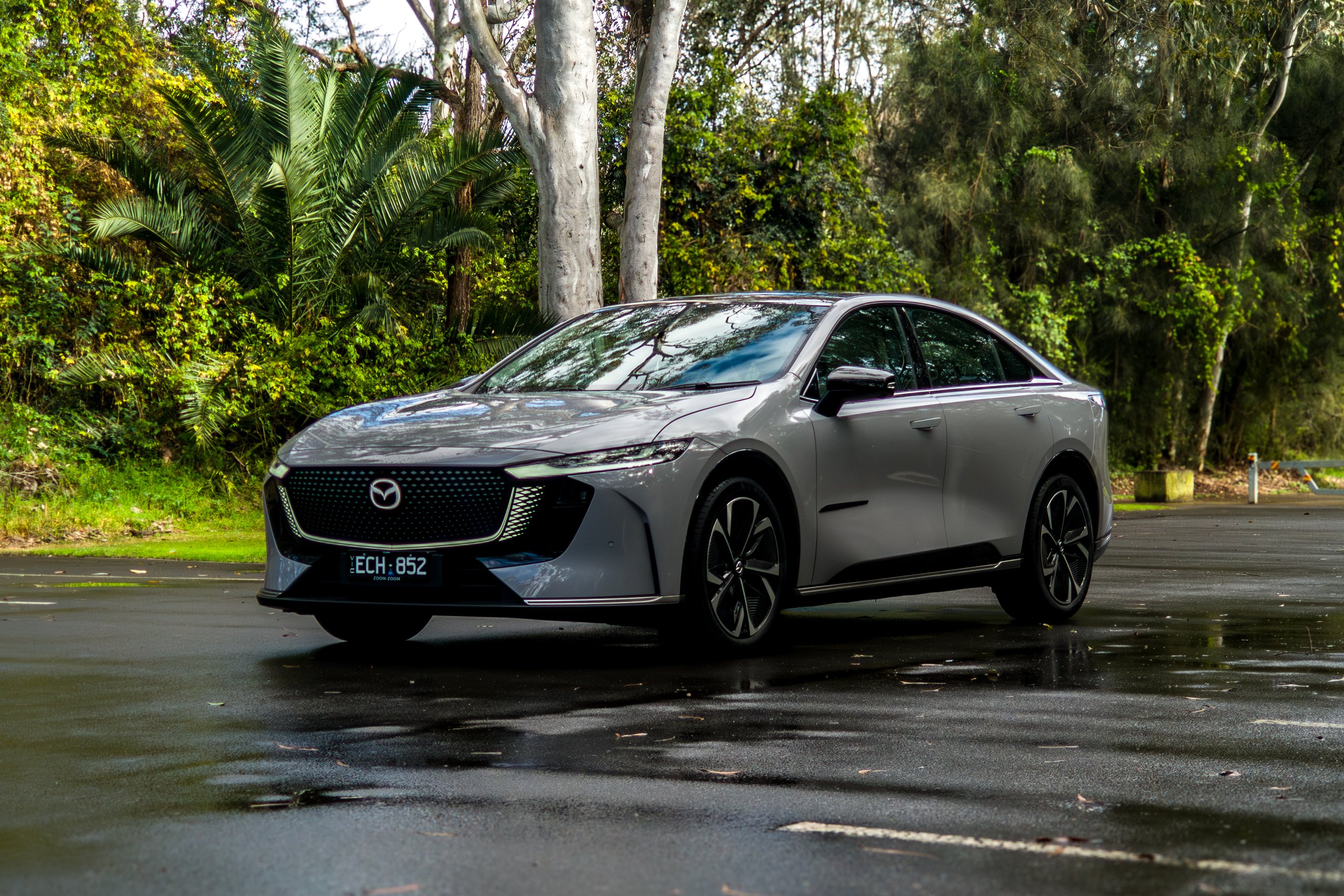 Light grey 2026 Mazda 6e GT parked on a wet road with palms and gum trees behind it