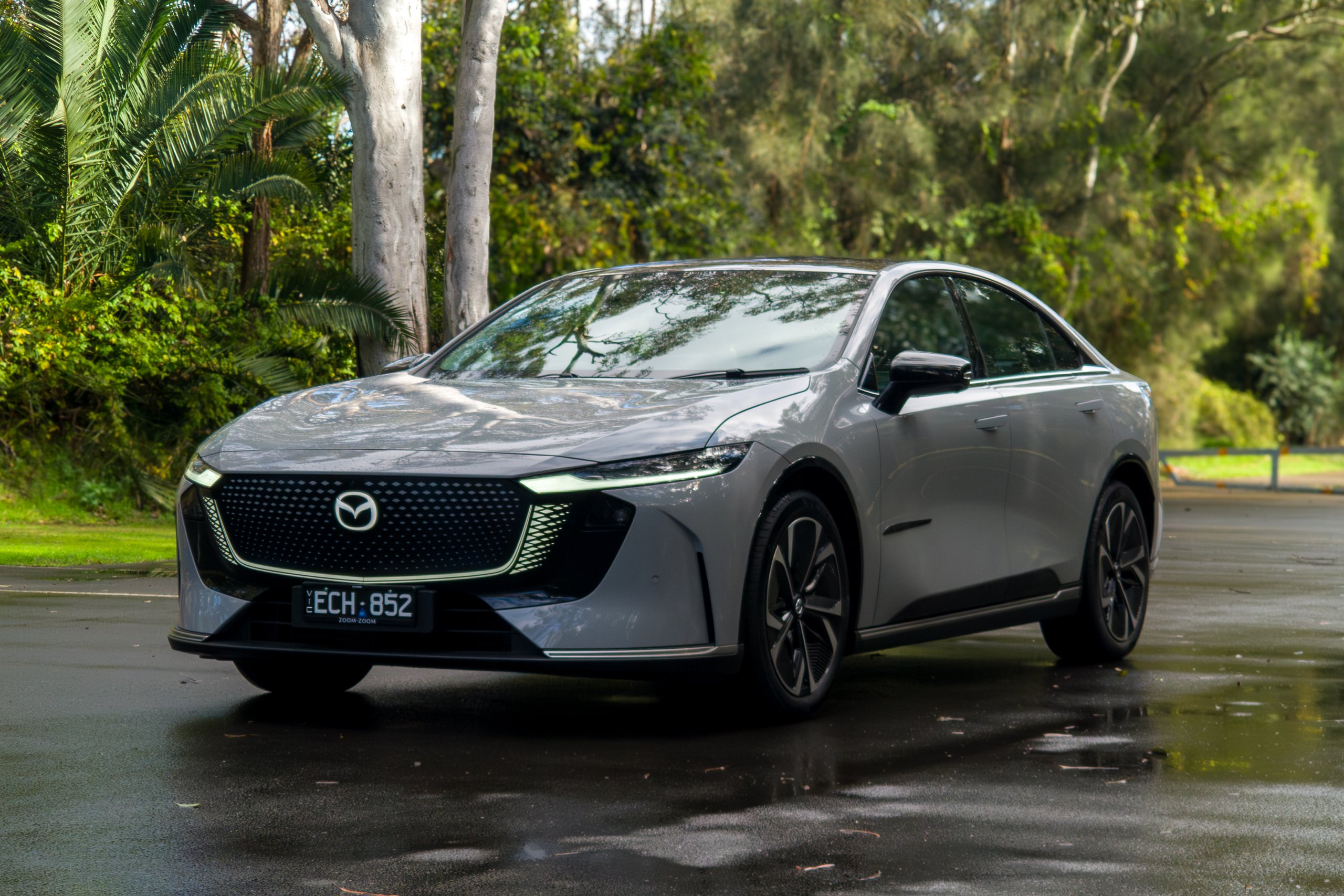 Light grey 2026 Mazda 6e GT electric sedan parked on wet bitumen, front three-quarter view