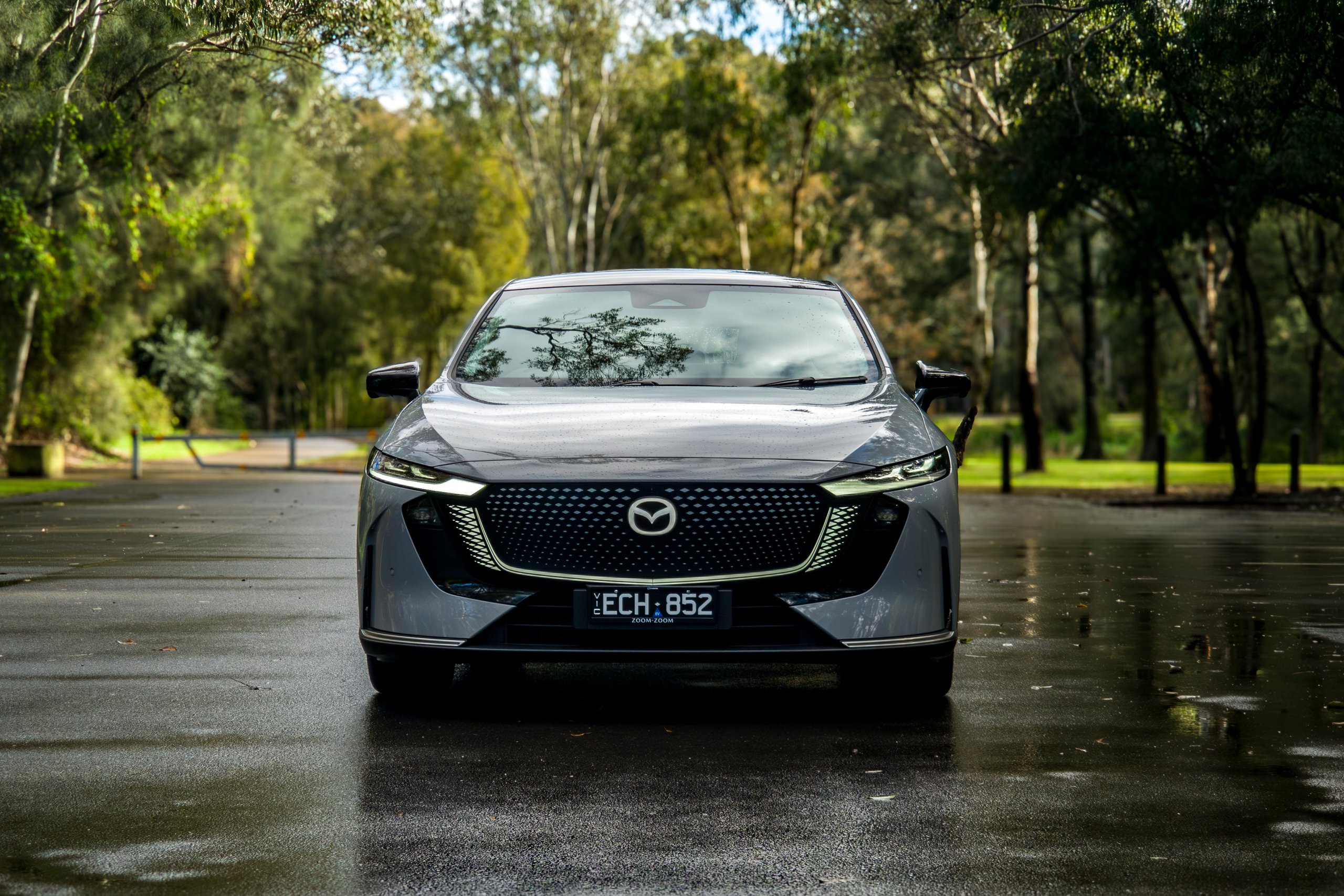 Head-on view of the light grey 2026 Mazda 6e GT parked on a wet forest road