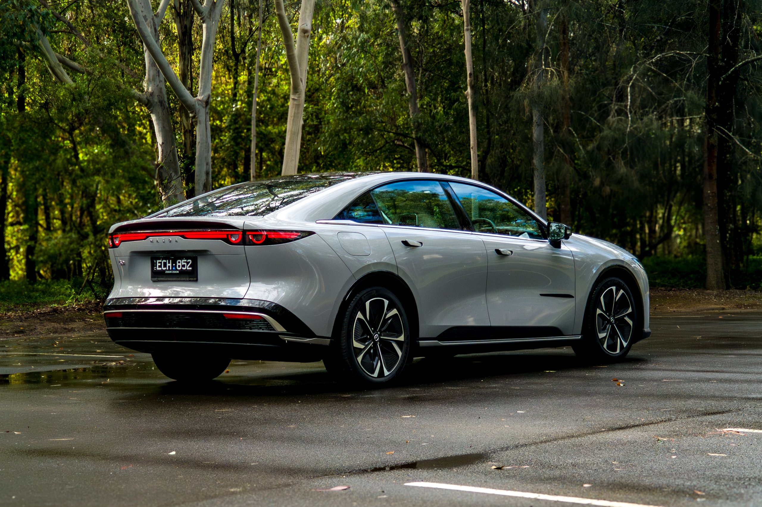 Rear three-quarter view of the light grey 2026 Mazda 6e GT on a wet bush road