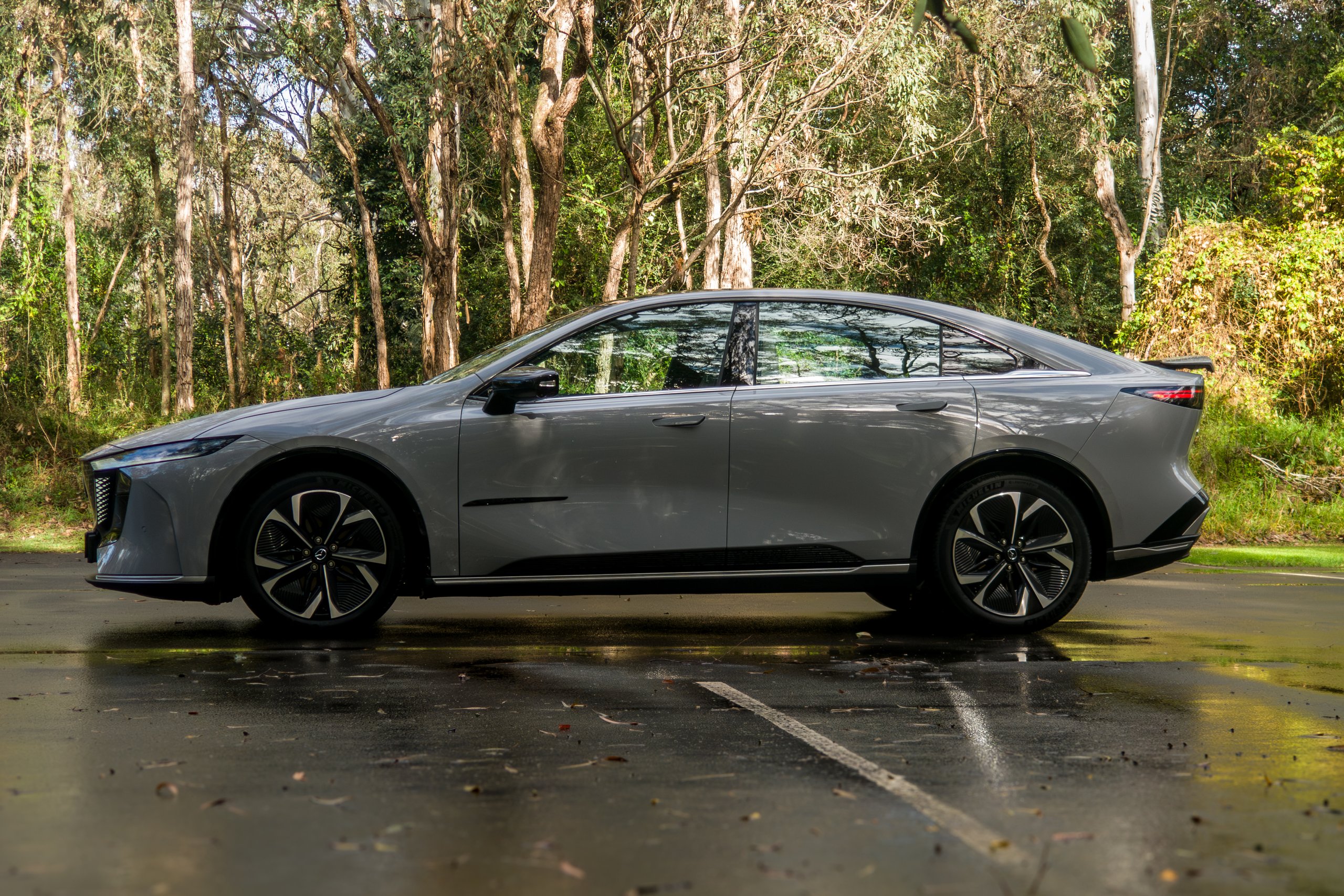 Passenger-side profile of the light grey 2026 Mazda 6e GT parked in front of gum trees