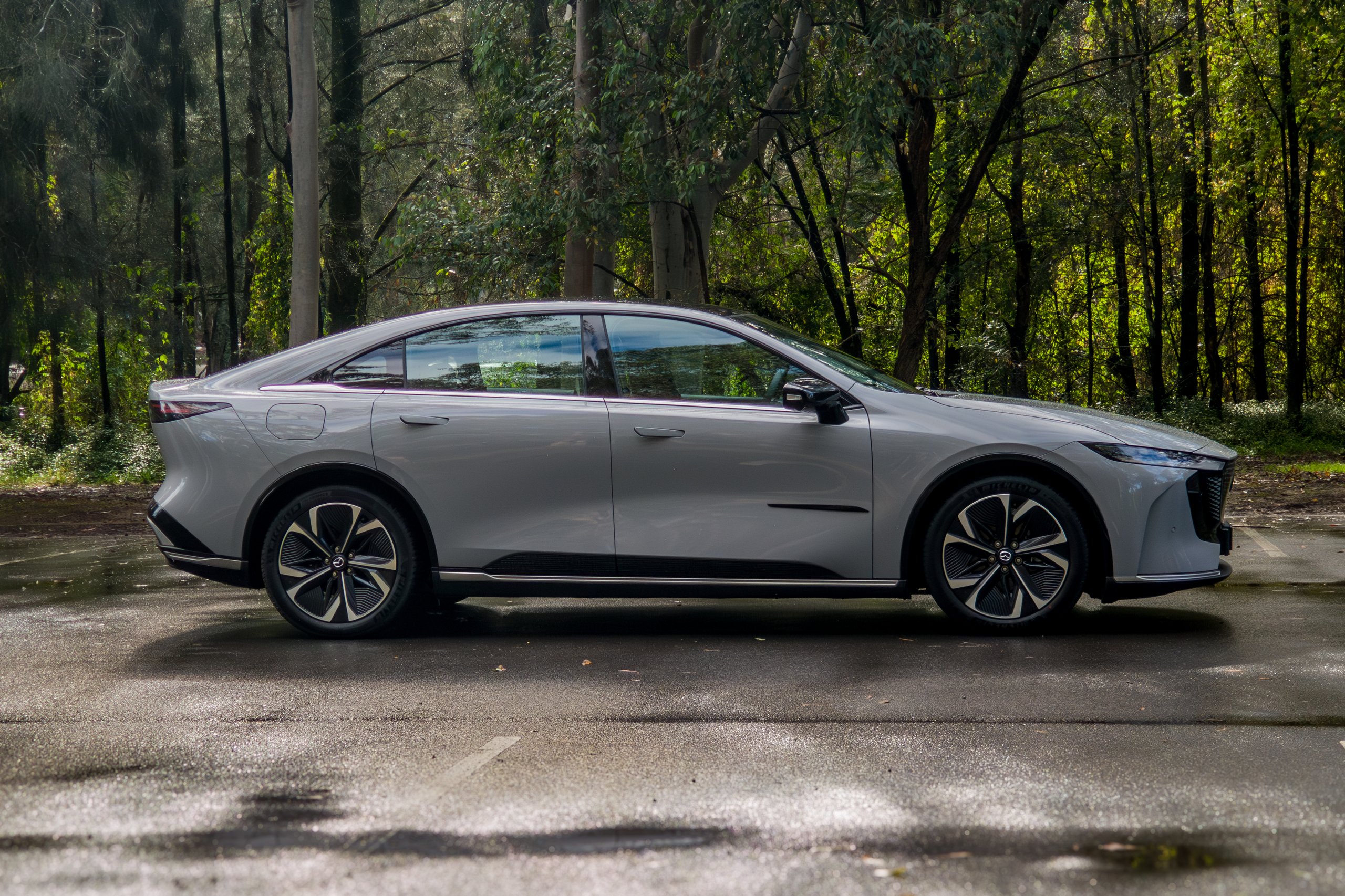 Full driver-side profile of the light grey 2026 Mazda 6e GT electric sedan on wet bitumen